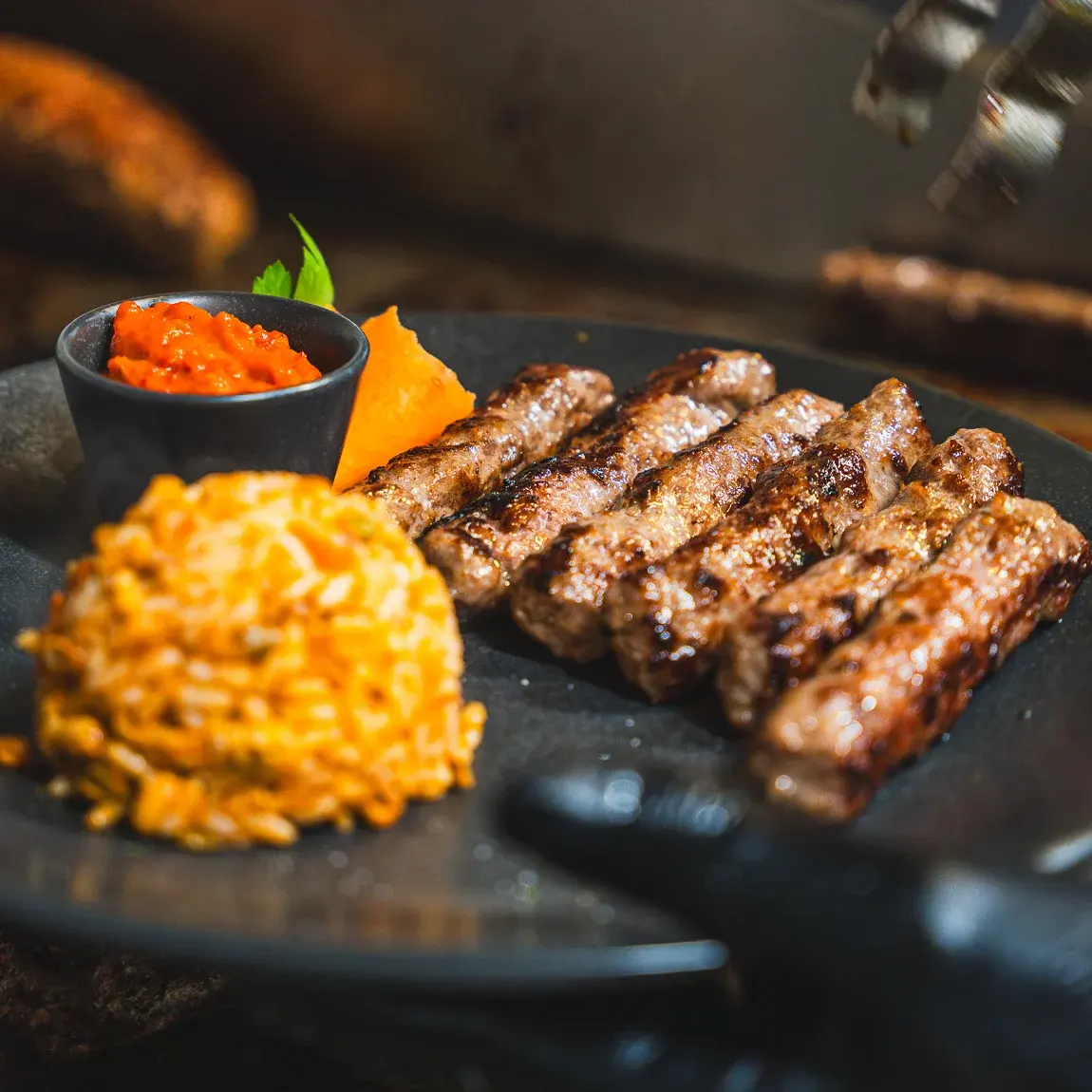Eine Reihe Cevapcici auf dunklem Teller, daneben Ajvar und Djuwetschreis