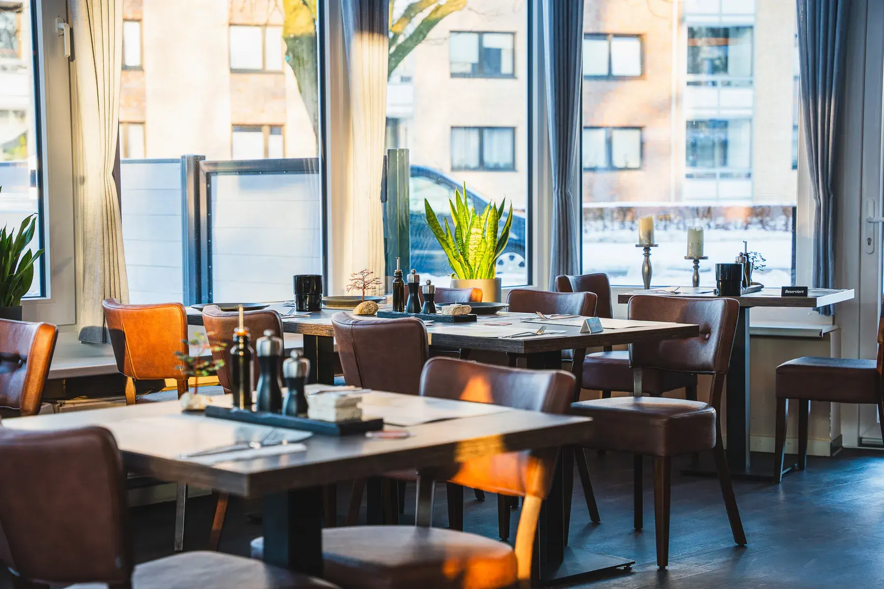 Der Wintergarten bei Tageslicht: gedeckte Tische und Lederstühle vor der raumhohen Fensterfront